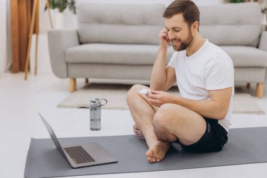 Çekici sakallı adam dizüstü bilgisayarın yanında kablosuz kulaklık tutuyor ve sabah egzersizi ya da yoga yapıyor..