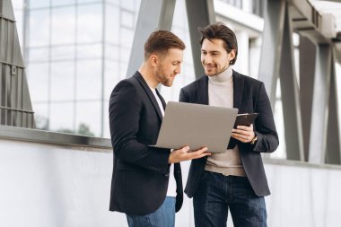 Takım elbiseli iki yakışıklı genç iş adamı bir dizüstü bilgisayar ve bir klasör tutuyorlar ve bir çalışma projesini tartışıyorlar ya da kentsel ofis binalarının arka planına karşı video görüşmesi yapıyorlar..