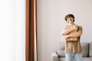 Alışveriş ve Teslimat. Kıvırcık Genç Müşteri, elinde karton kutuyla kameraya gülümsüyor. Heyecanlı Erkek Alıcı Sarılma Paketi. Alışveriş Konsepti