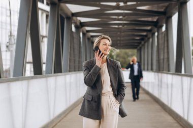 Takım elbiseli, gri ceketli, modern şehir ve ofis binalarında gülümseyen ve konuşan güzel bir iş kadınının portresi.