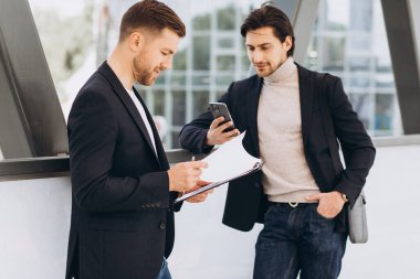 İki modern mutlu iş adamı ellerinde telefon ve dosya ile şehir ofislerinin ve binaların arka planına karşı bir şeyler tartışıyorlar.