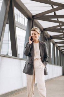 Takım elbiseli, gri ceketli, modern şehir ve ofis binalarında gülümseyen ve konuşan güzel bir iş kadınının portresi.