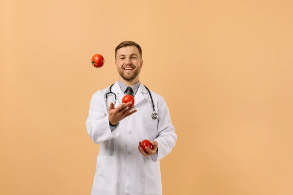 Męski Lekarz Dietetyk Stetoskopem Uśmiechający Się Żonglujący Pomidorami Beżowym Tle — Zdjęcie stockowe