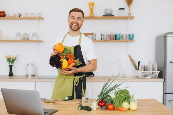 stock image Portrait of happy man at home, man cooking vegetable salad looking at camera and smiling, slicing vegetables, using laptop for online cooking training