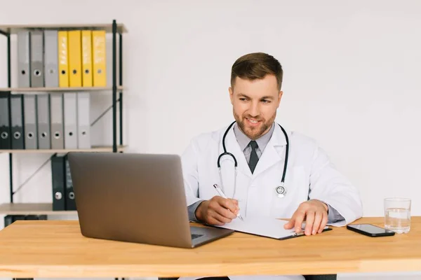職場でノートパソコンを持って座っている間 幸せな男性医師は オンライン医療のウェビナーやトレーニングセミナーを見て — ストック写真