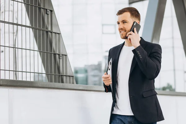 在城市建筑和办公室的背景下 穿着西服的快乐的现代男性商人拿着文件夹 通过电话交谈的画像 — 图库照片