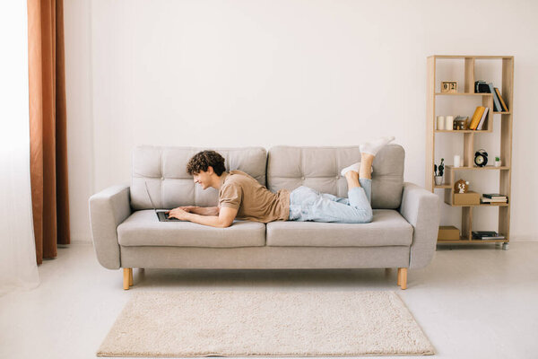 Portrait of attractive young curly hair man lying on the sofa and using laptop