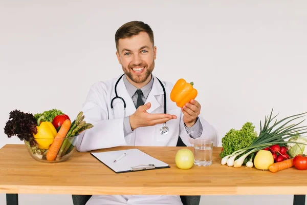 Happy doctor nutritionist sitting at workplace at desk in office among fresh vegetables and holding pepper, diet plan concept