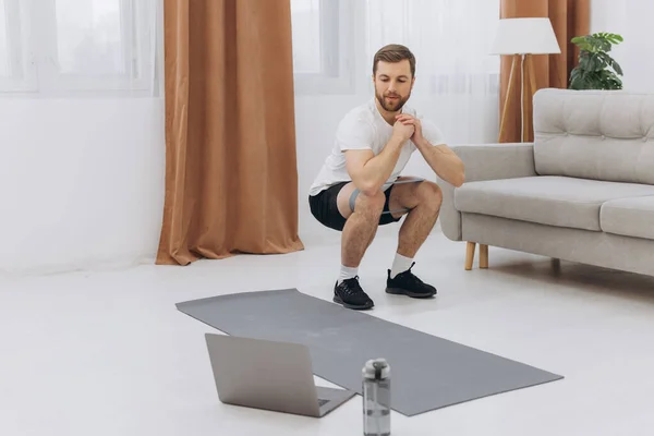 stock image Positive athletic bearded middle-aged man stretching at home, using laptop, watching sport videos on Internet, having fitness class online, copy space. Healthy lifestyle, sport on self-isolation