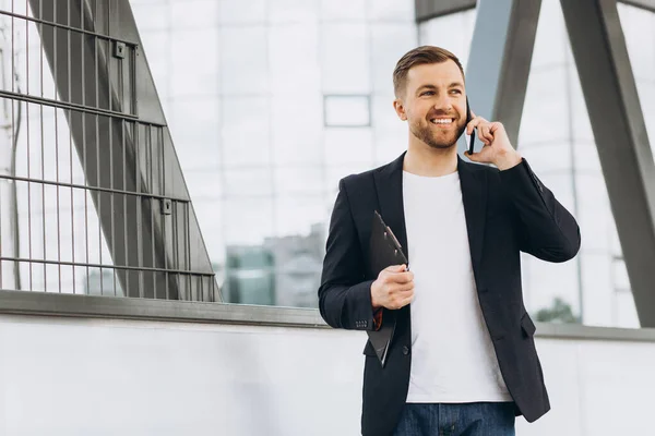 在城市建筑和办公室的背景下 穿着西服的快乐的现代男性商人拿着文件夹 通过电话交谈的画像 — 图库照片