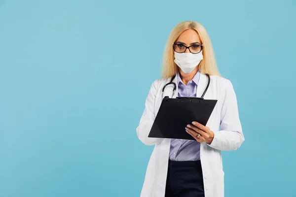 Mature woman doctor in mask holds folder on blue background. Covid-19 concept