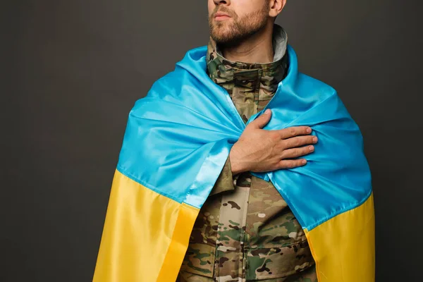 stock image Portrait of a courageous Ukrainian military man tied with the flag of Ukraine and holding hand on heart, freedom concept