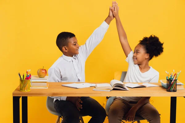 stock image Happy african american schoolgirl and mixed race schoolboy sitting together at desk and giving high five to each other on yellow background. Back to school concept.