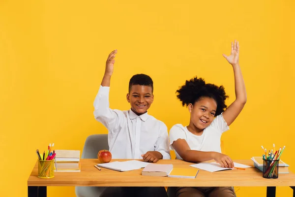 stock image Happy african american female schoolgirl and mixed race schoolboy sitting together at desk and stretching arms up on yellow background. Back to school concept.