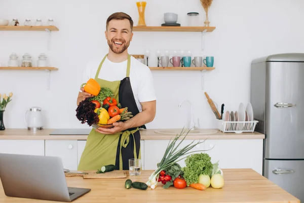 stock image Portrait of happy man at home, man cooking vegetable salad looking at camera and smiling, slicing vegetables, using laptop for online cooking training