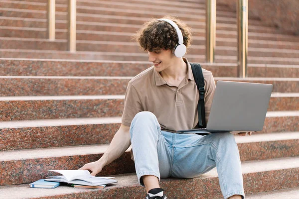 Concept of e-learning, distance study or remote learning concept, Young happy curly hair school guy, college or university student with backpack and headphones using laptop sitting on steps in university