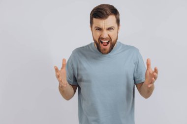 Mavi tişörtlü, modern sakallı orta yaşlı bir adamın portresi. Öfke duygusunu ve çığlıklarını beyaz arka planda gösteriyor.