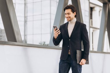 Takım elbiseli yakışıklı modern erkek işadamının portresi. Elinde evraklar var ve şehir binaları ve ofislerinin arka planında telefonla konuşuyor..