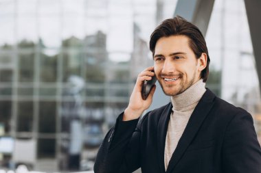 Takım elbiseli yakışıklı modern erkek işadamının portresi telefonla konuşuyor ve şehir binaları ve ofislerinin arka planında gülümsüyor..
