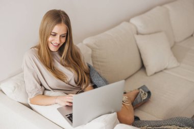 Pijamalı güzel sarışın kadın kanepede oturuyor ve dizüstü bilgisayarda çalışıyor, evden çalışma kavramı, karantina, yeni normal