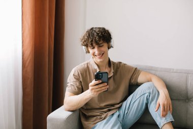 Oturma odasında kanepede oturup dinlenirken akıllı telefon kullanan kıvırcık saçlı genç adamın portresi.