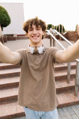 Kulaklıklı çekici kıvırcık erkek öğrencinin portresi gülümseyen ve selfie çeken, video çağrısı konsepti