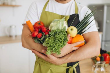 Evdeki mutfakta farklı taze sebzeler tutan mutlu bir adam.