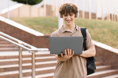 E-öğrenme, uzaktan öğrenme veya uzaktan öğrenme kavramı, üniversitede basamaklarda dizüstü bilgisayar kullanan genç kıvırcık saçlı okul çocuğu, sırt çantası ve kulaklıklı üniversite öğrencisi