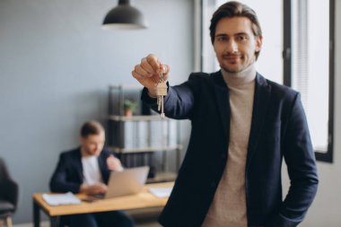 Takım elbiseli gülümseyen modern adam emlakçının ve ofisin arka planında satın alınan dairenin anahtarlarını tutuyor.