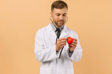 Erkek doktor kardiyolog bej arka planda steteskopla kalbi dinliyor.