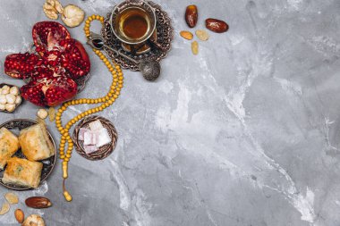 Geleneksel Arap sofra takımı ve yemek takımlarıyla süslü afiş, bir fincan çay, Kuran ve gri taşlı arka planda tespih. Ramazan masa üstü görünümü