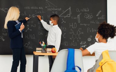 Bayan öğretmen okulda tahtaya yakın çocuklara ders veriyor. Karantina, maske modu, covid-19. Okula geri dön..