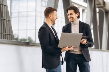 Takım elbiseli iki yakışıklı genç iş adamı bir dizüstü bilgisayar ve bir klasör tutuyorlar ve bir çalışma projesini tartışıyorlar ya da kentsel ofis binalarının arka planına karşı video görüşmesi yapıyorlar..
