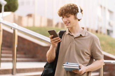 Sırt çantası ve kitaplarıyla üniversitenin yanındaki telefona bakan mutlu ve çekici öğrenci.