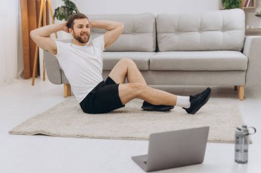 Pozitif atletik sakallı orta yaşlı bir adam evde esniyor, dizüstü bilgisayar kullanıyor, internette spor videoları izliyor, fitness dersi alıyor, kopya çekiyor. Sağlıklı yaşam tarzı, kendini soyutlama sporu.