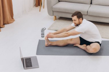 Pozitif atletik sakallı orta yaşlı bir adam evde esniyor, dizüstü bilgisayar kullanıyor, internette spor videoları izliyor, fitness dersi alıyor, kopya çekiyor. Sağlıklı yaşam tarzı, kendini soyutlama sporu.