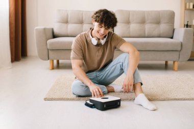 Çekici genç kıvırcık adam, evdeki oturma odasında halı temizlerken temizlik robotunu çalıştırıyor..