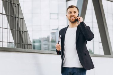 Takım elbiseli mutlu modern erkek işadamının portresi elinde belgelerle şehir binaları ve ofislerinin arka planında telefonla konuşuyor..