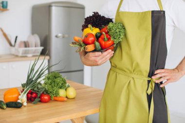 Mutfağın arka planında taze sebze tabağı ve çeşitli sebzelerle dolu mutfak masası tutan bir adam..