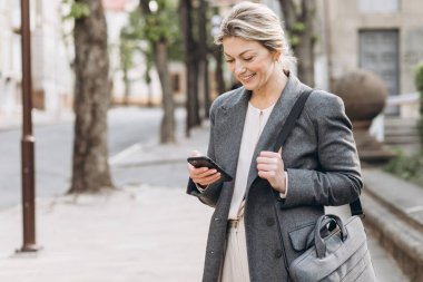 Gri ceketli olgun sarışın bir iş kadını. Gülümsüyor ve elinde telefonla şehirde dolaşıyor.