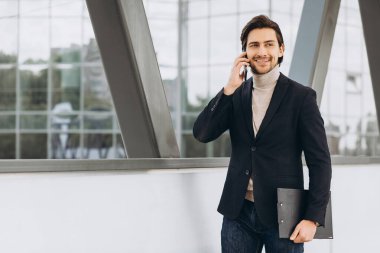 Takım elbiseli yakışıklı modern erkek işadamının portresi. Elinde evraklar var ve şehir binaları ve ofislerinin arka planında telefonla konuşuyor..