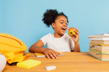Mutlu Afro-Amerikan liseli kız sınıfta oturuyor ve mavi arka planda elmayla öğle yemeği yiyor. Okul konseptine dönelim..