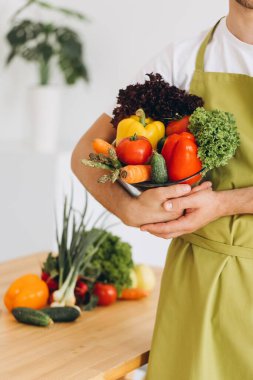Mutfağın arka planında taze sebze tabağı ve çeşitli sebzelerle dolu mutfak masası tutan bir adam..