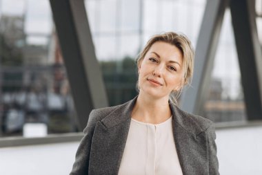 Yetişkin bir iş kadınının portresi modern şehir ve ofis binalarının arka planında duygularla gülümsüyor.