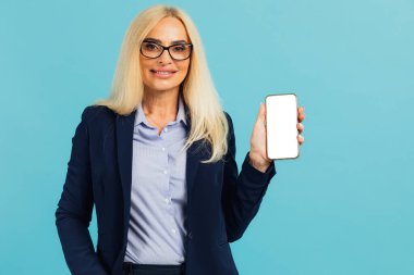 Resmi takım elbiseli, gülümseyen olgun bir iş kadınının portresi. Elinde beyaz, boş bir cihaz ekranı olan akıllı bir telefon, kameraya yakın, mavi stüdyo. Model için boş alanı olan Gadget