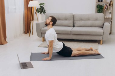 Pozitif atletik sakallı orta yaşlı bir adam evde esniyor, dizüstü bilgisayar kullanıyor, internette spor videoları izliyor, fitness dersi alıyor, kopya çekiyor. Sağlıklı yaşam tarzı, kendini soyutlama sporu.