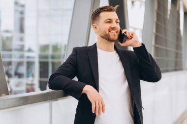 Takım elbiseli yakışıklı erkek işadamının portresi şehir binaları ve ofislerinin arka planında telefonla konuşuyor..