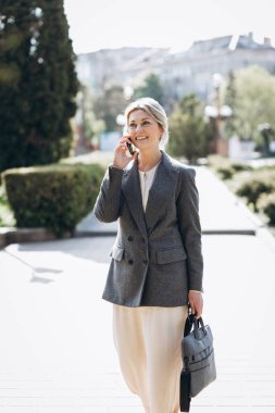 Olgun sarışın gülümseyen iş kadını şehrin sokaklarında yürüyor ve telefonda konuşuyor.