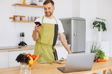 Çekici genç adam mutfakta dizüstü bilgisayarla yemek pişiriyor ve akıllı telefondan konuşuyor..