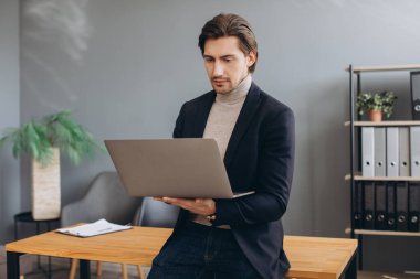 Modern ofiste takım elbiseli ve dizüstü bilgisayar kullanan genç ve neşeli iş adamının görüntüsü